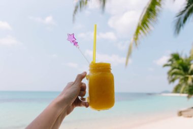 Güneşli bir sahil sahnesi, sahil kenarında palmiye ağaçları ve nazik dalgalarla çevriliyken canlı bir tropikal içecekten keyif alan birini gösterir..