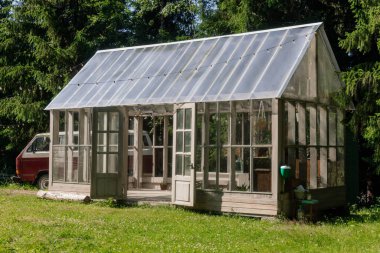 Bu kırsal serada, berrak mavi gökyüzünün altındaki yüksek ağaçlarla çevrili sakin bir bahçede kurulu ahşap bir çerçeve ve cam paneller bulunuyor..