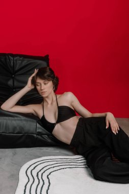 Modern kapalı bir ortamda, siyah elbiseli bir kadın zarif bir şekilde şık bir sandalyede poz verir. Kalın kırmızı arka plan, onun rahat tavırlarını ve modaya uygun tarzını vurguluyor..