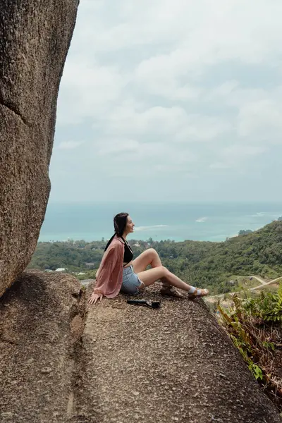 Uçsuz bucaksız bir okyanusa bakan kayalık bir uçurumda oturan bir kadın. Önünde yeşil yapraklar ve yukarıda da puslu mavi bir gökyüzü var..