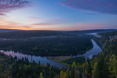 Huzurlu bir gündoğumu gökyüzünde yumuşak renkler bırakır, yemyeşil ormanların arasından akan kıvrımlı bir nehri aydınlatarak, dingin bir sabah atmosferi yaratır..