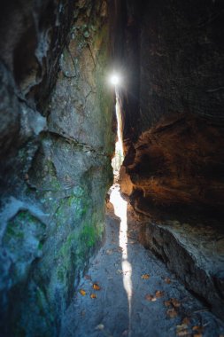 Dar kayalık, düz taş duvarlı bir geçit güneş ışığının içeri akmasına izin verir, kumlu zemini aydınlatırken etrafındaki yapraklar da sakin bir atmosfer yaratır..