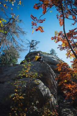 Bir dağcı, berrak mavi gökyüzünün altında parlak sonbahar yapraklarıyla süslenmiş büyük bir kaya oluşumunu ustalıkla yükselerek doğanın barışçıl bir ortamda güzelliğini gösterir..