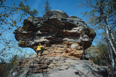 Sarı ceket giyen bir yürüyüşçü, açık mavi gökyüzünün altında ağaçlarla çevrili etkileyici bir kaya formasyonunun yanında poz verir ve bölgenin doğal cazibesini vurgular..