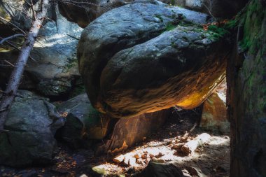 Öğleden sonra güneşi, sessiz bir ormanda iki taş oluşumu arasında tehlikeli bir şekilde dengelenmiş devasa bir kayayı aydınlatır. Çevreleyen yeşillik huzur ortamına derinlik katıyor.