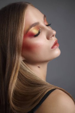 Model, başını eğerek zarifçe poz veriyor. Kırmızı ve sarı tonlarda çarpıcı bir makyaj sergiliyor. Yumuşak stüdyo ışıkları altında yüz hatlarını geliştiriyor..