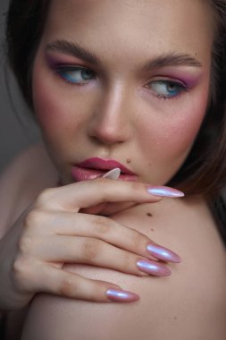 Renkli göz makyajı ve yumuşak allık ile tamamlanmış parlak bir kaplamayla çarpıcı uzun tırnaklar sergiliyor. Duruşu, manikür ve makyajının sanatını ön plana çıkarıyor..