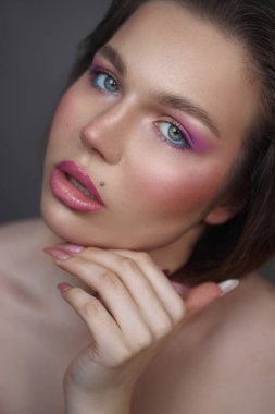 Kadın eli yüzünün yanında zarif bir şekilde poz veriyor, mor ve pembe tonlarda renkli makyaj yapıyor. yumuşak ışıklandırma onun çarpıcı özelliklerini ve pürüzsüz cildini vurgular.