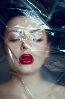 Cesur kırmızı dudaklı ve sanatsal göz makyajlı bir kadın yüzü yarı saydam naylonla kaplanmış şekilde poz veriyor..