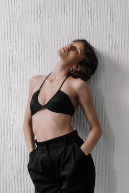 Genç bir kadın dokulu bir duvara karşı poz verirken kendine güvenir. Şık bir siyah bikini bluzu ve terzi pantolonu giyiyor, rahat bir pozda modern ve şık bir estetik taşıyor..