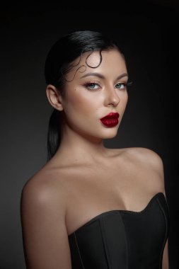 Kadın kendine güvenerek poz veriyor çarpıcı bir makyajla koyu kırmızı dudaklar ve tanımlanmış kirpiklerle. Parlak saç stili, yumuşak, karanlık bir atmosferde genel zarafete katkı sağlıyor..