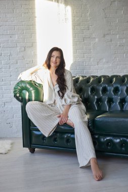 Kadın, öğleden sonra güneş ışığıyla vurgulanan parlak bir odada, yeşil bir kanepede rahatça oturuyor. Şık bir elbise giymiş ve rahat ve rahat görünüyor..