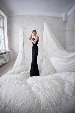 Model zarif bir şekilde siyah bir elbisenin içinde duruyor, iyi ışıklandırılmış bir iç mekanda çarpıcı bir kontrastlık yaratan eşsiz beyaz kumaş biçimleriyle çevrili. stil entelektüellik ve sanatsallık yayar..