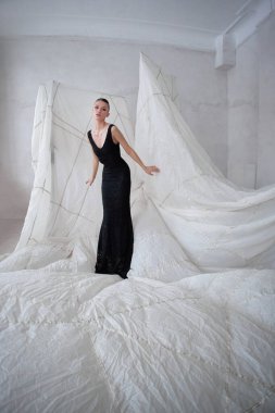 Minimalist bir stüdyo ortamında siyah bir elbise giyen manken zarif pozlar veriyor. Arkaplan büyük soyut beyaz kumaş şekillerinden oluşur, çarpıcı bir kontrast oluşturur.