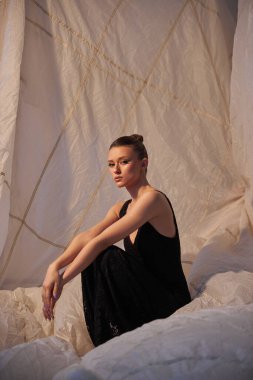 Model, minimalist bir stüdyoda beyaz bir kumaş zemin üzerinde zarif bir şekilde oturur. Etrafındaki yumuşak dokularla çelişen siyah bir kıyafet giyiyor, dingin bir güzelliğin görüntüsünü yakalıyor..