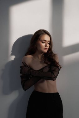 Bir duvarın yanında zarif bir şekilde duran genç bir kadın, vücudunu vurgulayan saf kumaşla örtülmüş. Güneş ışığı yumuşak gölgeler yaratır, kapalı mekan atmosferinin sanatsal atmosferini güçlendirir..