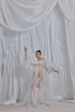 Model, beyaz kumaşın önünde zarif bir poz veren, dramatik kolları olan şeffaf bir elbise sergiliyor. giysi tasarımını vurgulamak ruhani ve sanatsal bir duygu..