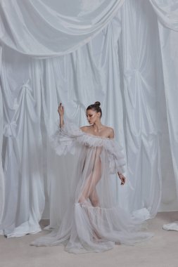 Model havadar bir ortamda duruyor. Etrafında zarif bir şekilde süzülen ince kumaşlar içinde. yumuşak kumaşlar sanatsal moda gösterisine rüya gibi bir kalite ekler..