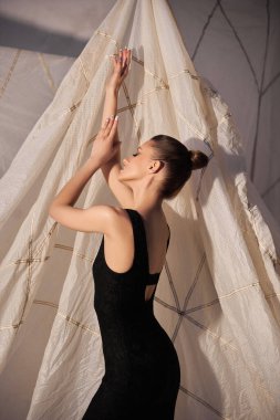 Model, kolları dalgalı bir kumaşın yanında zarif bir şekilde poz veren parlak siyah bir elbise sergiliyor. onun siluetini vurgulayan yumuşak ışıklandırma özellikleri ayarlamak.