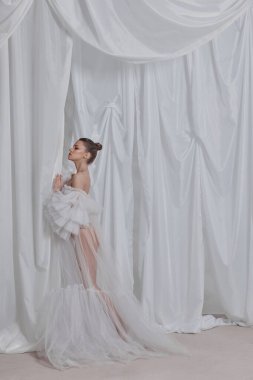 Modeller çarpıcı beyaz bir elbisenin içinde yumuşak kumaşlı bir arka plana yaslanarak zarifçe duruyorlar. Moda çekimi sırasında hassas kumaş ve modellerin zarif pozlarını ön plana çıkarıyor..