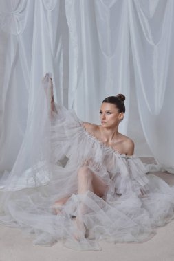 Zarif bir saç stiline sahip bir model zarif bir şekilde oturuyor, katıksız bir elbise giyiyor. yumuşak, örtülü arka plan sakin bir atmosfer yaratıyor, narin kumaşı ve özgüvenli ifadesini vurguluyor..