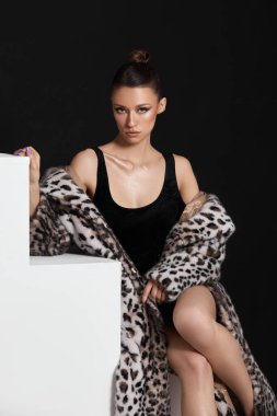 Şık bir model, leopar desenli kürk manto ile tamamlanmış siyah bir elbise sergiliyor. Geceleri hafif ışıklandırmayla aydınlatılan minimalist bir stüdyoda zerafet ve güven yayıyor..