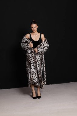 Model, kendine güvenen bir şekilde stüdyoda duruyor, siyah bir kıyafetin üzerine şık bir leopar desenli ceket giyiyor. Siyah arkaplan, bu fotoğraf çekiminde yapılan moda açıklamasını vurguluyor.