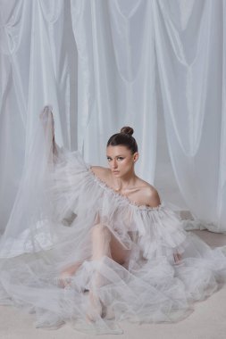 Model yumuşak bir zeminde pürüzlü kolları olan şeffaf bir sabahlıkla zarifçe oturuyor. Arkadaki yumuşak, örtülü kumaş rüya gibi bir atmosfer yaratıyor..