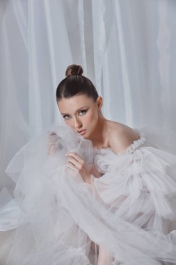 Model, yumuşak bir ifadeyle düşünceli bir poz veren narin bir beyaz tül kıyafeti sergiliyor. Kapalı alan arka planı, kumaşın yarattığı ruhani atmosferi güçlendirir..