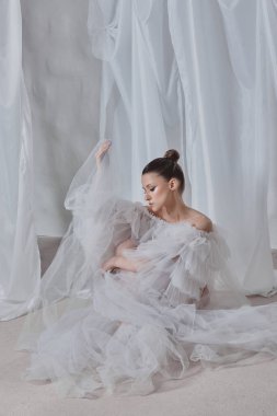 Narin beyaz kumaşlı bir model yumuşak bir yüzey üzerinde zarifçe poz veriyor, katıksız perdelerle çevrili..