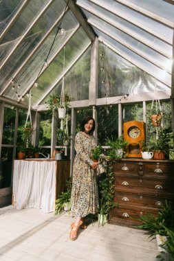 Çiçekli elbiseli bir kadın, canlı bir serada taze bitkiler düzenlerken gülümsüyor. Cam duvarlardan geçen güneş ışığı filtreleri, çeşitli saksı bitkilerini ve klasik dekorları vurguluyor..