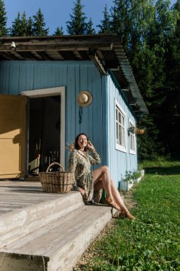 Bir kadın sıcak bir öğleden sonra güneşlenirken, büyüleyici mavi bir kulübenin verandasında oturuyor. Etrafı ağaçlarla çevrili, bir sepet tutuyor, görünüşte rahat ve doğadan memnun..