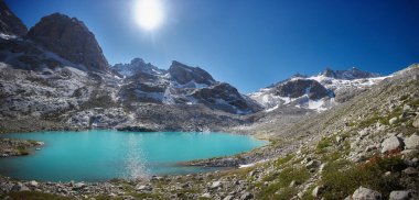 Canlı turkuaz göl, parlak güneş ışığını yansıtan görkemli dağların arasında oturur. engebeli arazi ve berrak gökyüzü, doğayı keşfetmek ve keyif almak için ideal, çarpıcı bir doğal manzara yaratır..