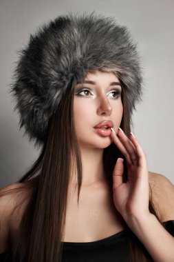 Genç bir kadın, içeride poz verirken düşünceli bir bakışla şık gri bir kürk şapka sergiliyor. Onun çarpıcı makyajı ve uzun saçı yakalanan anın modaya uygun cazibesini artırıyor..