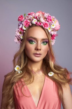 Genç bir kadın, pembe ve beyaz çiçeklerden yapılmış çiçek başlığıyla stüdyoda poz veriyor. Makyajında canlı renkler var. Şık bir elbise giyiyor. İlkbahar zarafetinin vücut bulmuş hâli..