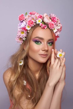 Mor ve yeşilliklerle süslü renkli çiçek tacıyla süslenmiş canlı makyajlar sergiliyor. Elleriyle zarif pozlar veriyor, saçlarında narin çiçeklerle..