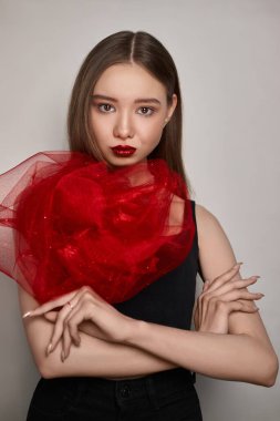 Model, minimalist bir stüdyoda kendine güvenen bir moda ifadesi olarak dramatik bir kırmızı eşarp sergiliyor. Makyajı özelliklerini vurgulayarak büyüleyici bir görsel karşıtlık yaratıyor..