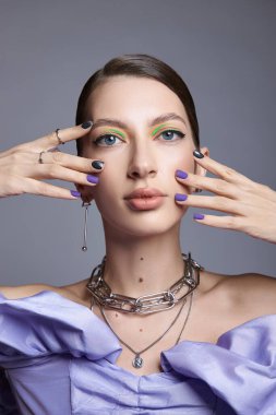 Kadın kendine güvenen bir şekilde stüdyoda poz veriyor, çarpıcı makyajını neon aksanı ve kalın tırnak renkleriyle vurguluyor. Şık elbisesini tamamlayan katmanlı mücevherler takıyor..