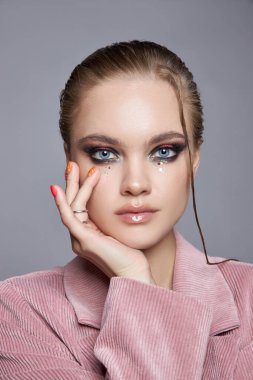 Model bir stüdyoda zarif pozlar veriyor, benzersiz makyaj yapıyor cesur gözler ve parlak dudaklar sergiliyor. Pembe bir ceket giyer ve kendine güveni tamdır..