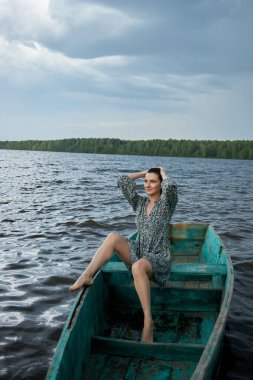 Kadın sakin bir gölde, mavi bir teknede, arka planda bulutlu bir gökyüzünün altında bir orman belirirken sudaki huzurun tadını çıkarıyor..