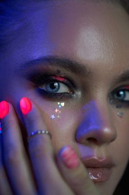 Çarpıcı mavi gözleri ve parlak neon tırnakları olan genç bir kadın yüzüyle etkileşime giriyor. Renkli bir gecede ışıltılı ve ışıltılı detaylar içeren eşsiz bir makyaj bakışını vurguluyor..