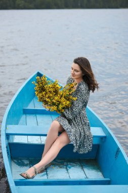 Kadın sakin göl sularında, doğa tarafından çevrili mavi bir teknede dinleniyor. Sıcak bir öğleden sonra huzurlu bir anın tadını çıkaran canlı bir buket sarı çiçek tutuyor..