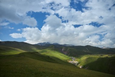 Geniş yeşil tepeler, dağlık bir bölgede bulutlarla dolu canlı bir gökyüzünün altında uzanır ve sakin bir manzarada gündüz saatlerinde doğanın güzelliğini gösterir..