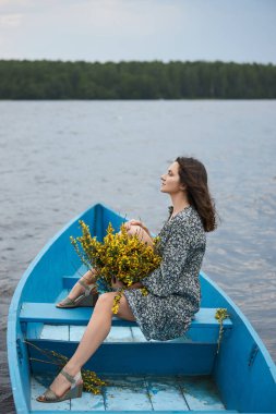 Genç bir kadın sakin bir suda mavi bir teknede otururken, parlak bir buket sarı çiçeği tutarken düşünceli bir şekilde ileriye bakarken huzurlu bir an yaşanıyor. Bereketli ağaçlar uzak kıyıya sınır çiziyor..