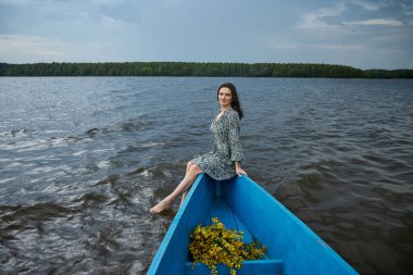 Kadın zarif bir şekilde mavi bir teknede oturuyor, ayakları suyun içinde. Şablonlu bir elbise giyiyor ve dalgalı dalgaların ve bulutlu bir gökyüzünün altında yemyeşil bir kıyı şeridinin ortasında rahatlamış görünüyor..