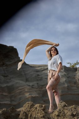 Kumlu bir tepede duran bir kadın bulutlu bir gökyüzünün altındaki kayalık kayalıklara atkısını neşeyle kaldırır. Rüzgâr saçlarıyla oynuyor o anı kucaklarken.