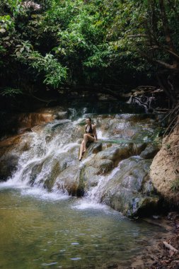 Kadın, tropikal bir ormandaki yoğun yeşilliklerle çevrili kayalıklarda oturmuş şelalenin yanında huzurlu bir anın tadını çıkarıyor. Güneş ışığı yaprakların arasından süzülerek barışçıl bir ortam yaratır..