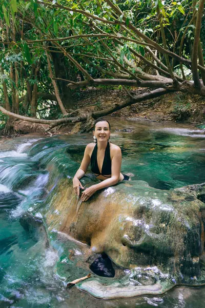 Genç bir kadın, canlı tropikal bitki örtüsüyle çevrili, açık bir doğal baharda dinlenir. Bu sakin ortamda sakin su ve güzel kayalarla meşgul olurken gülümsüyor..