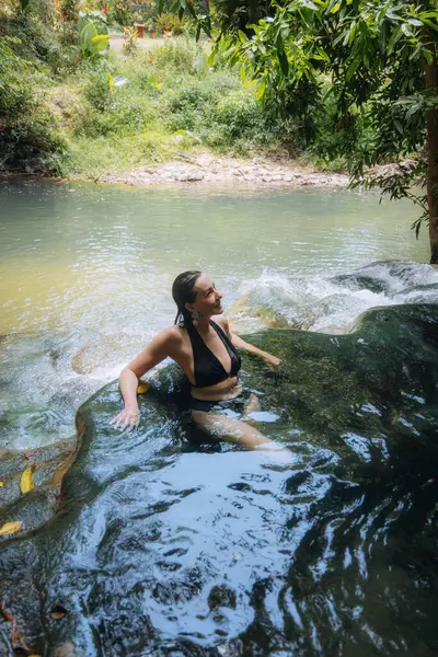 Kadın berrak, doğal bir havuzda, canlı yeşil yapraklarla çevrili bir şekilde otururken huzurlu bir anın tadını çıkarıyor. Huzurlu su, ılık güneşin altındaki kayaların üzerinden yavaşça akar..