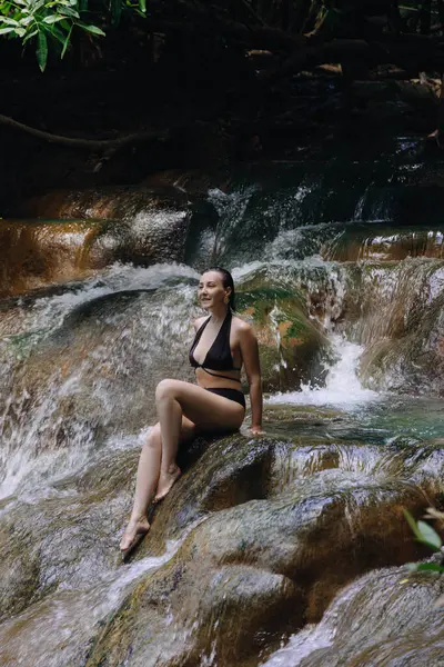 Siyah mayo giymiş bir kadın, canlı bir tropikal ortamda akan bir şelalenin yanındaki pürüzsüz bir kayanın üzerinde dinlenir ve güneşli bir günde doğanın sakinliğini gösterir..